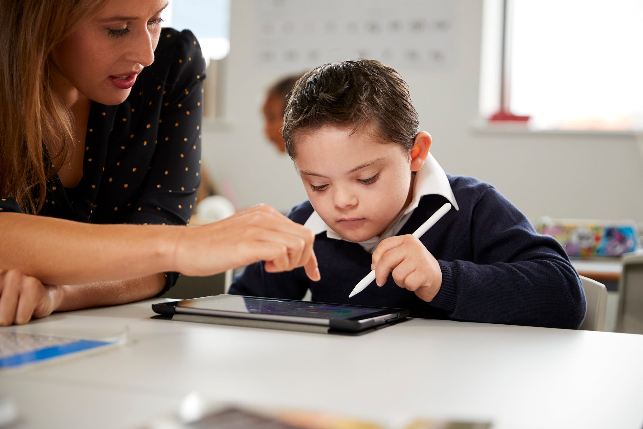 Young female teacher working with a Down syndrome schoolboy sitting at desk using a tablet computer in a classroom