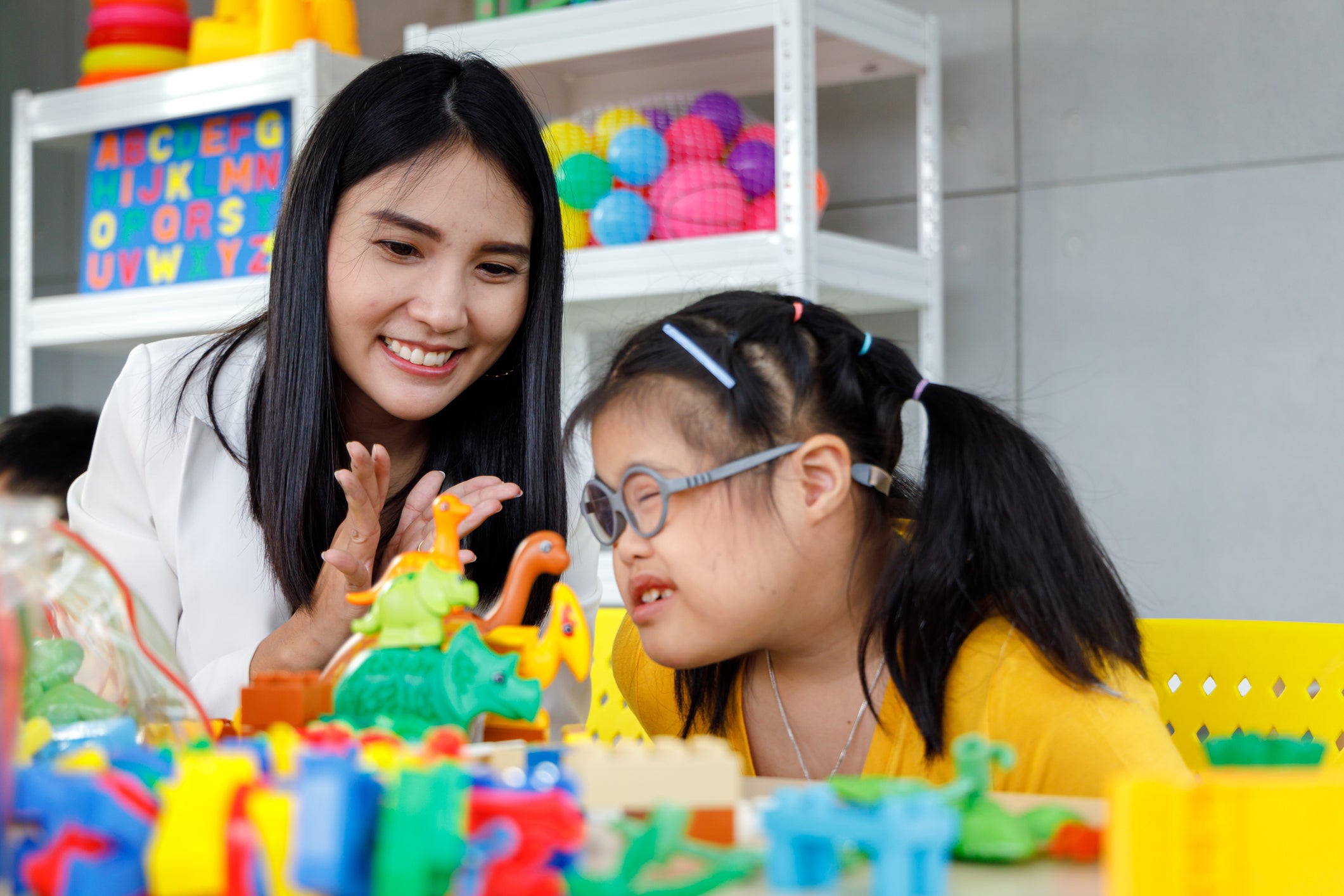 Smiling Asian woman in white blazer sitting with girl with Down's syndrome playing with colorful plastic dinosaur toy.