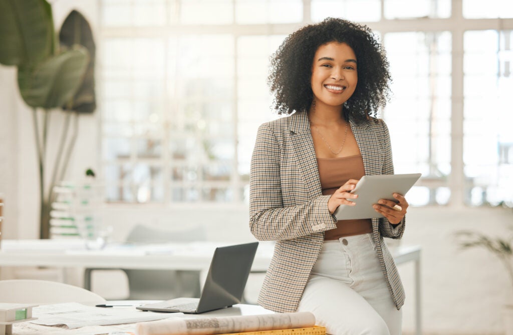 Black woman with curly shoulder length hair is smiling and standing in her office using a tablet