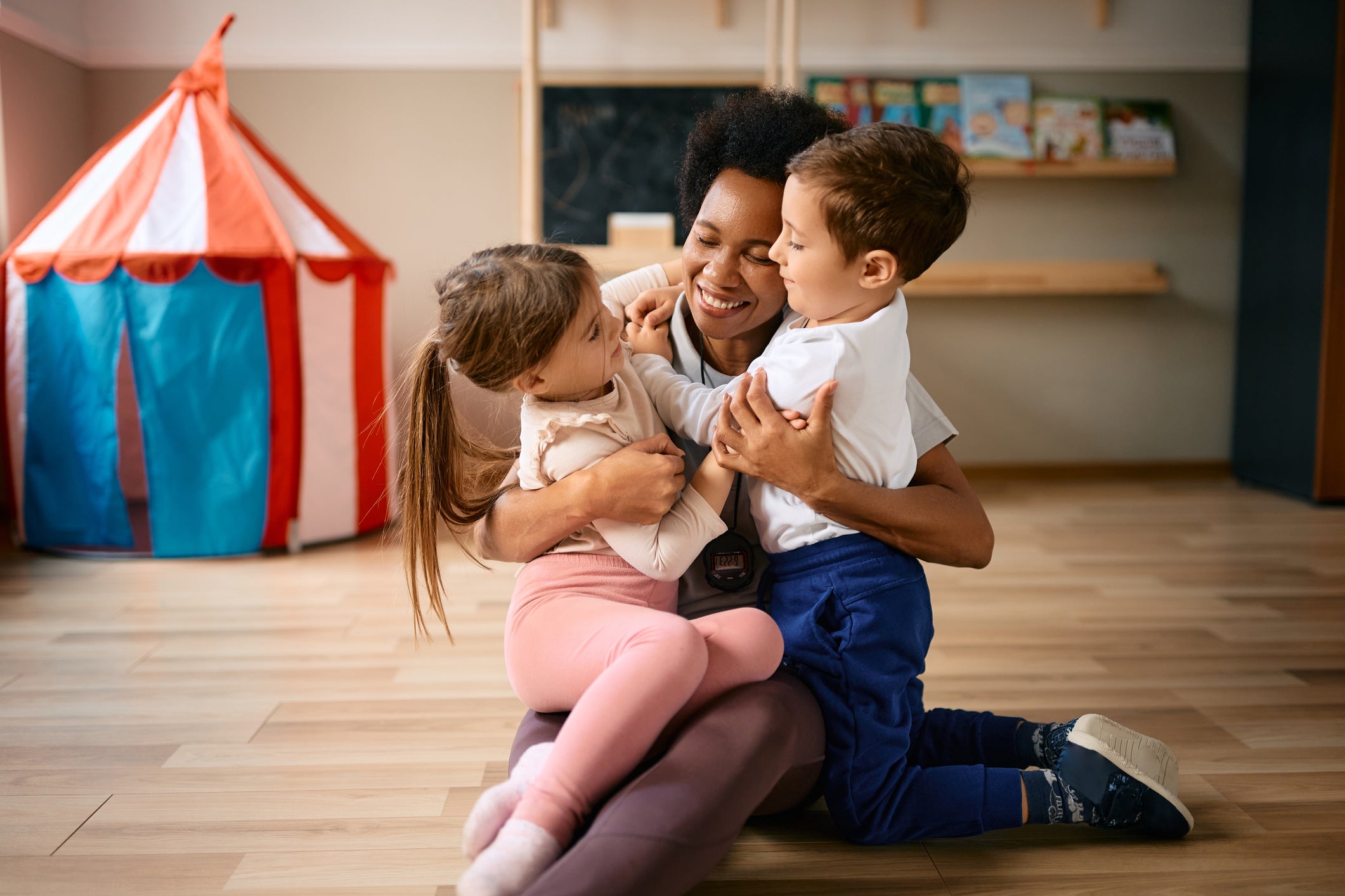 Black teacher embracing two Latino children - one girl wearing pink leggings and white t-shirt and one boy wearing blue pants and white t-shirt on the wooden floor of a classroom