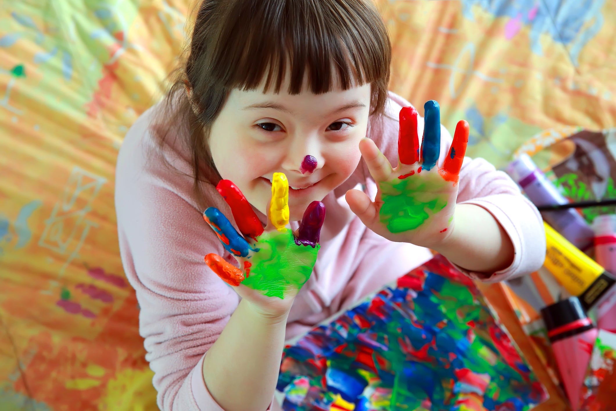 Little girl looking up with dark brown hair in bangs and painted hands with palms up. She is standing on a multicolored sheet.