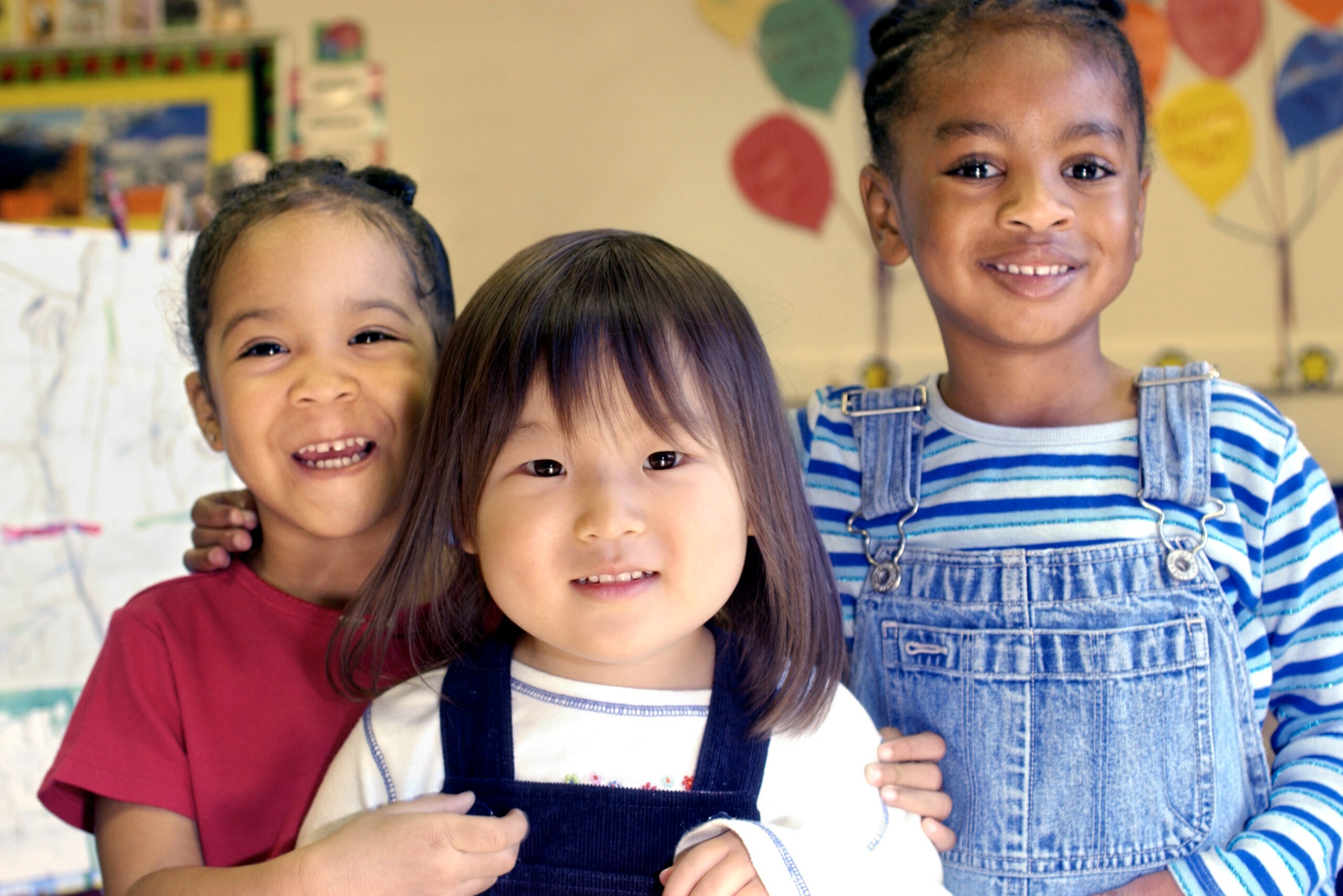 One Latina girl with braided pulled back black hair and red shirt standing next to an Asian girl with dark brown/black hair wearing a white t-shirt and dark blue overalls standing next to a black girl with a blue/white striped t-shirt and blue jean overalls in a classroom.
