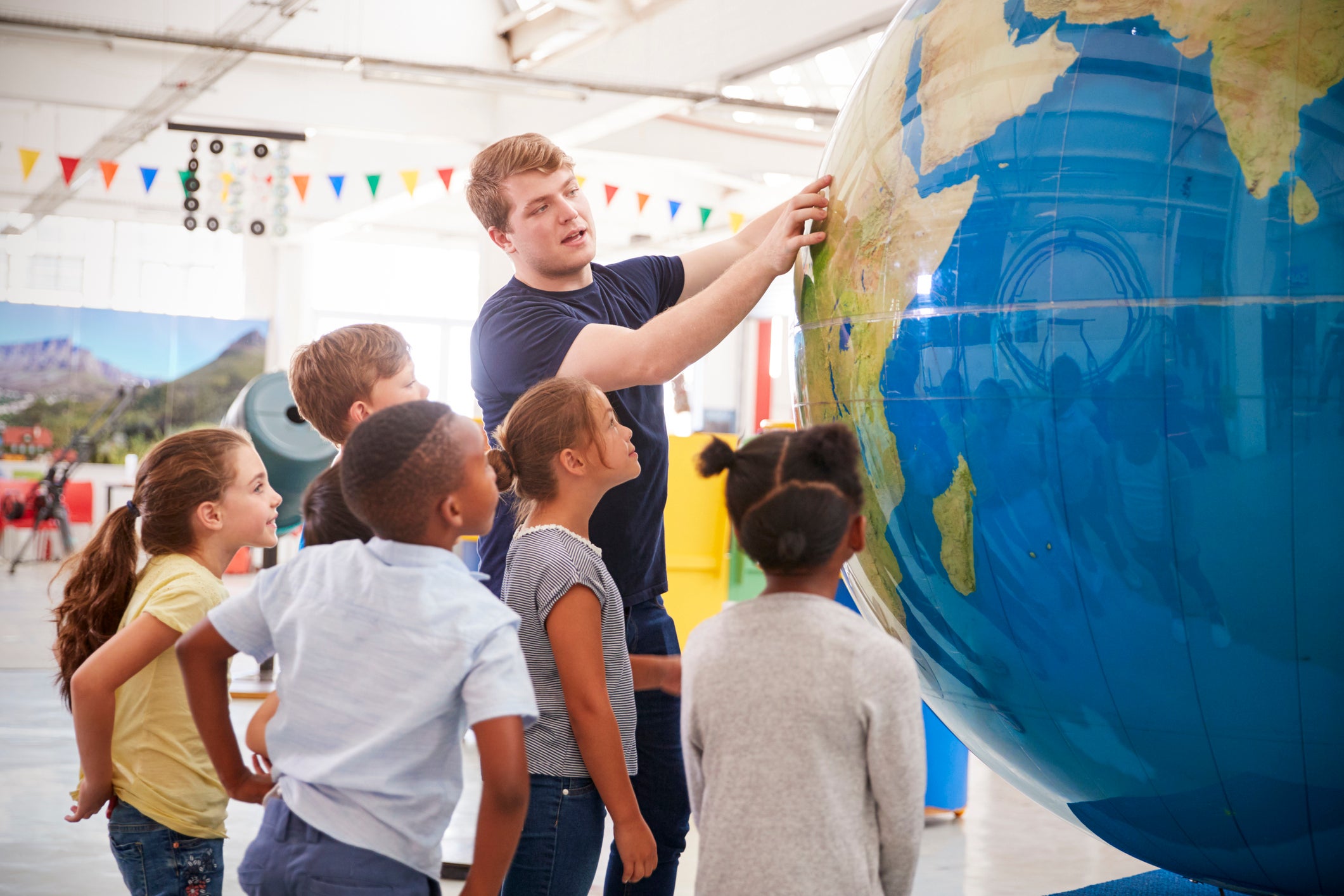 Tall white man with brown hair in blue t-shirt and blue jeans points to South America on a large inflated globe. Kids watch giant globe