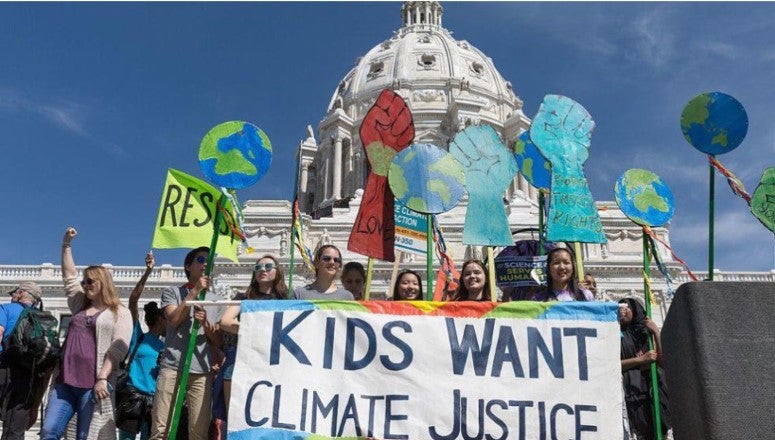 13 young adults of differing genders and ethnicities with raised fists and posters of the world globe, right clenched fists, and a banner reading 'Kids Want Climate Justice' standing in front of the White House Capitol building